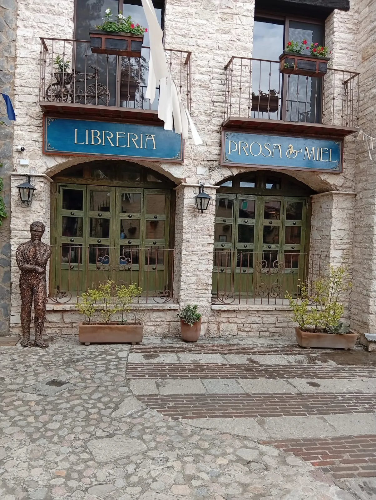 Fachada de la librería Prosa & Miel en Val'Quirico: dos letreros azules de madera sobre puertas verdes, muros de piedra y calle empedrada.