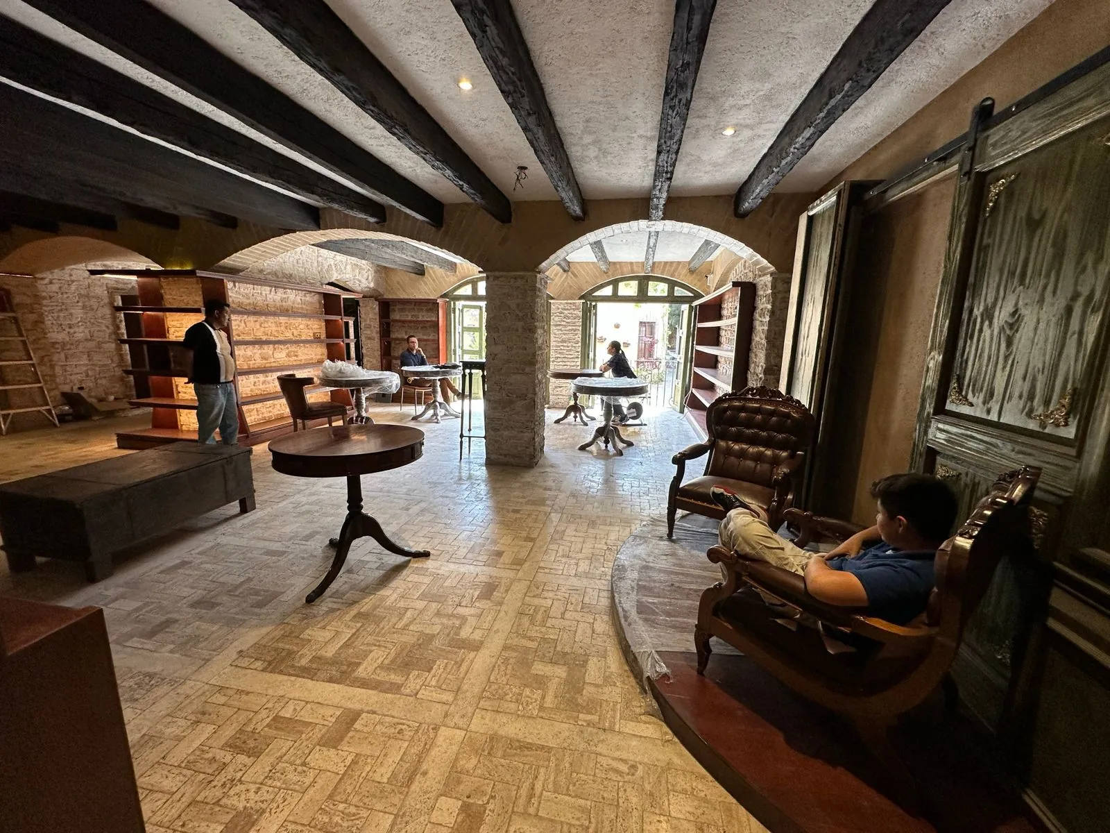 Sala interior con vigas negras, arcos de piedra, mesas redondas de madera y sillones de piel.